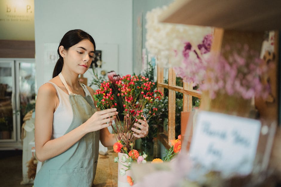 A young woman with dark hair tied back, wearing a light-colored apron over a sleeveless top, stands in a flower shop holding a vibrant bouquet of small, round, pink and red flowers with green stems. T