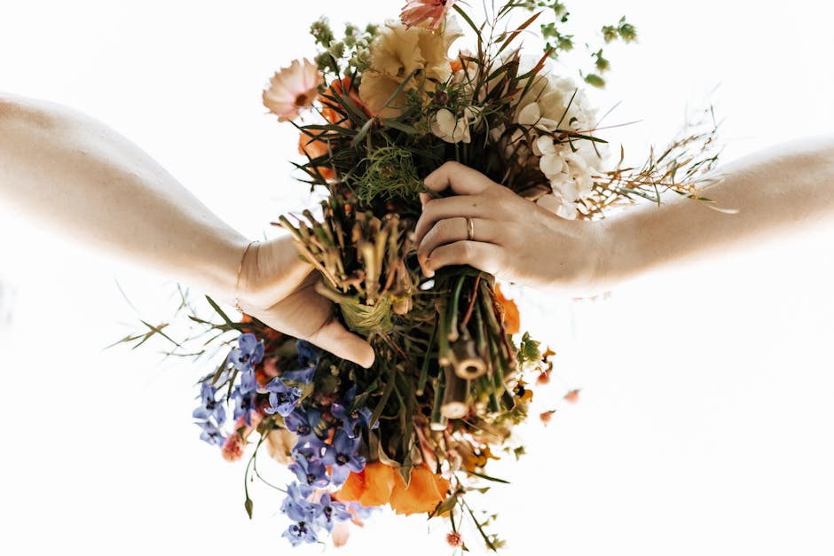 A pair of hands holding a colorful bouquet of fresh flowers, featuring orange, white, and purple blooms, wrapped in simple green stems and tied with a natural twine. The bouquet includes a mix of rose