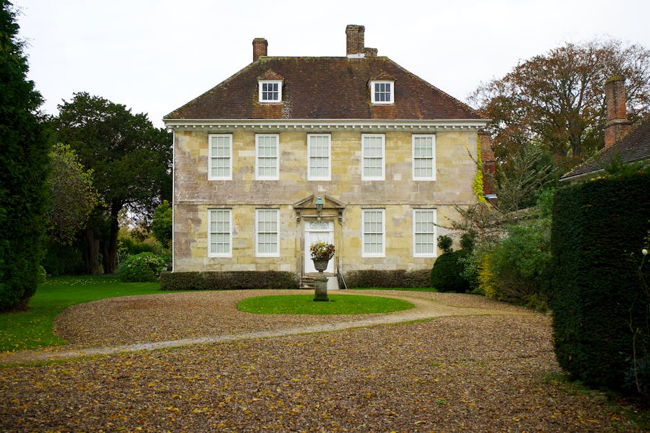A stately, three-storey stone mansion with a steeply pitched, reddish-brown tiled roof and multiple chimneys, set amidst lush green trees and well-manicured gardens. The building features symmetrical