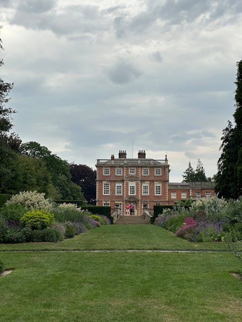 The image depicts a historic brick manor house set amidst a lush, well-manicured garden with a straight central lawn flanked by colorful flower borders. The garden features a variety of flowering plan
