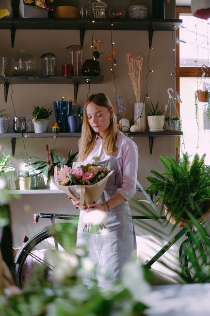 A young woman with long, light brown hair is holding a bouquet of fresh flowers wrapped in brown paper, featuring pink roses, white lilies, and greenery. She is dressed in a white shirt and apron, sta