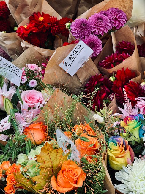 A vibrant display of freshly cut flowers arranged in brown paper wrapping at a floral shop. The bouquet includes bright red dahlias, lavender-purple chrysanthemums, soft pink roses, and orange roses, 