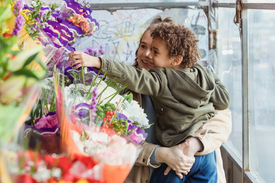 A young boy with curly hair and a smiling woman, possibly his mother, are inside a flower shop, surrounded by a vibrant display of fresh flowers in various colors including pink, white, purple, and re