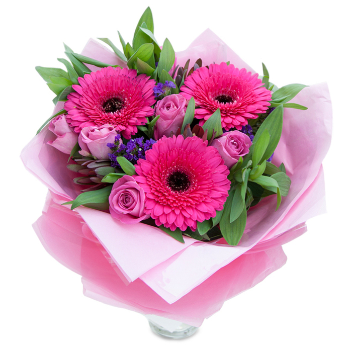 There is a quiet, garden-like charm to this arrangement that balances playfulness with polish, a composition an attentive Crayford florist might place by a sunlit window. Three exuberant fuchsia Gerbera daisies make the visual statement, their broad petals opening like small fireworks around distinctive dark centres that anchor the eye. Interspersed are softly tinted lavender-pink roses in early bloom, their edges slightly rolled inward in a way that speaks of freshness and slow, graceful opening. Scattered throughout are sprigs of purple statice-tiny, textured clusters that introduce a cool, almost blue-leaning contrast against the warm pinks and lend the bouquet a hint of wildflower spontaneity. The greenery is generous and purposeful: glossy salal-like leaves and slender eucalyptus stems weave through the flowers, giving the arrangement a structured, living backdrop and a faint herbal fragrance that complements the sweeter rose scent. Presentation is deliberate: a pale pink tissue inner wrap cushions the stems while a translucent, crinkled fuchsia-pink cellophane layer supplies a subtle glint, the whole package folded with care so it reads as both intimate and celebratory. The top rim of a clear glass vase is visible at the bouquet's base, suggesting ready-to-display freshness. This piece suits occasions that call for warmth and affection-birthdays, anniversary surprises, or a thoughtful 'just because'-and is especially fitting for deliveries around Crayford's high street, local residences, and nearby communities in Bexley, arriving bright and composed.